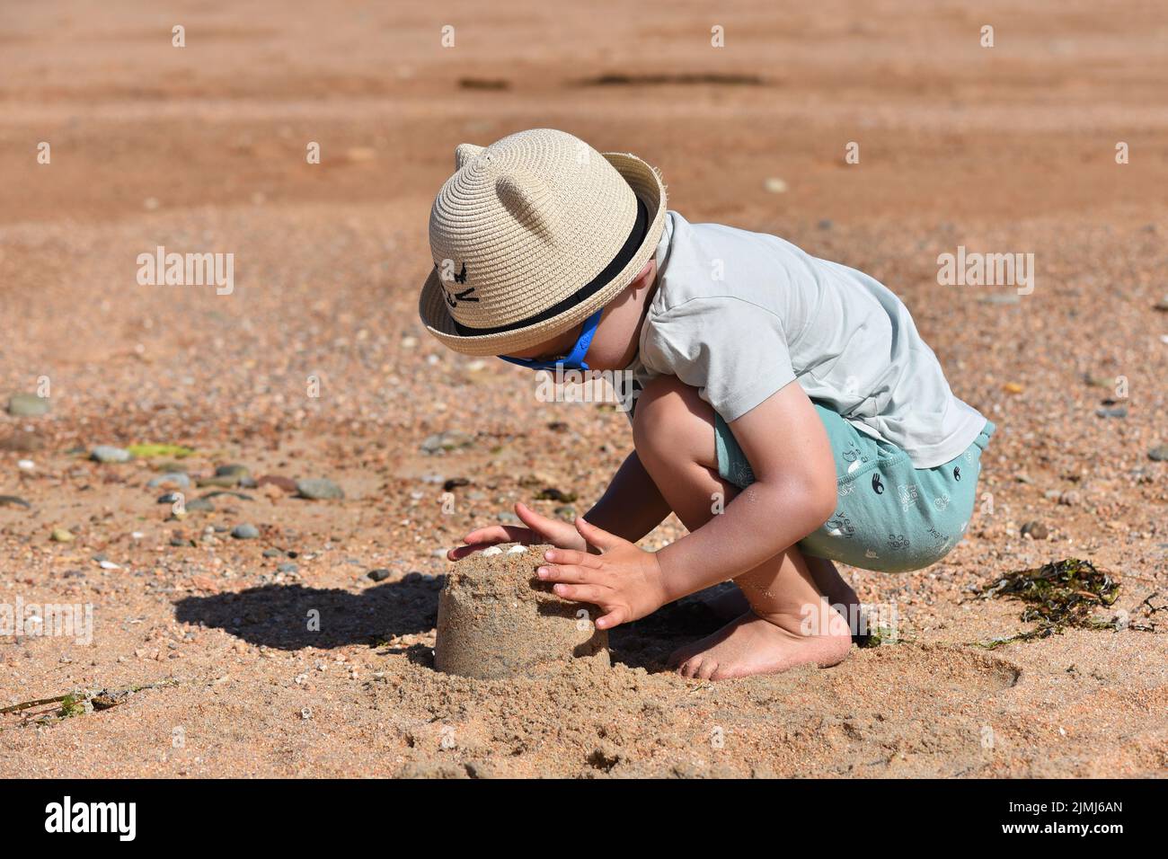 Young crouched child wearing a nice hat on a sunny beach and building a sandcastle decorated with seashells. Stock Photo