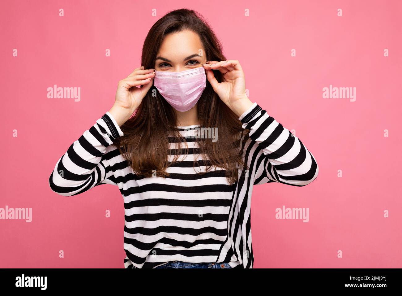 Photo shot of young beautiful brunette woman in reusable virus protective mask on face against coronavirus isolated on the pink Stock Photo