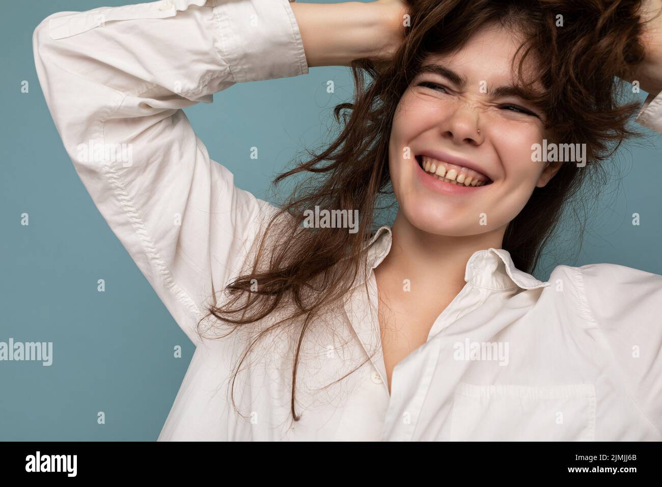 Portrait of beautiful positive cheerful cute smiling young brunette woman in casual white shirt isolated on blue background with Stock Photo