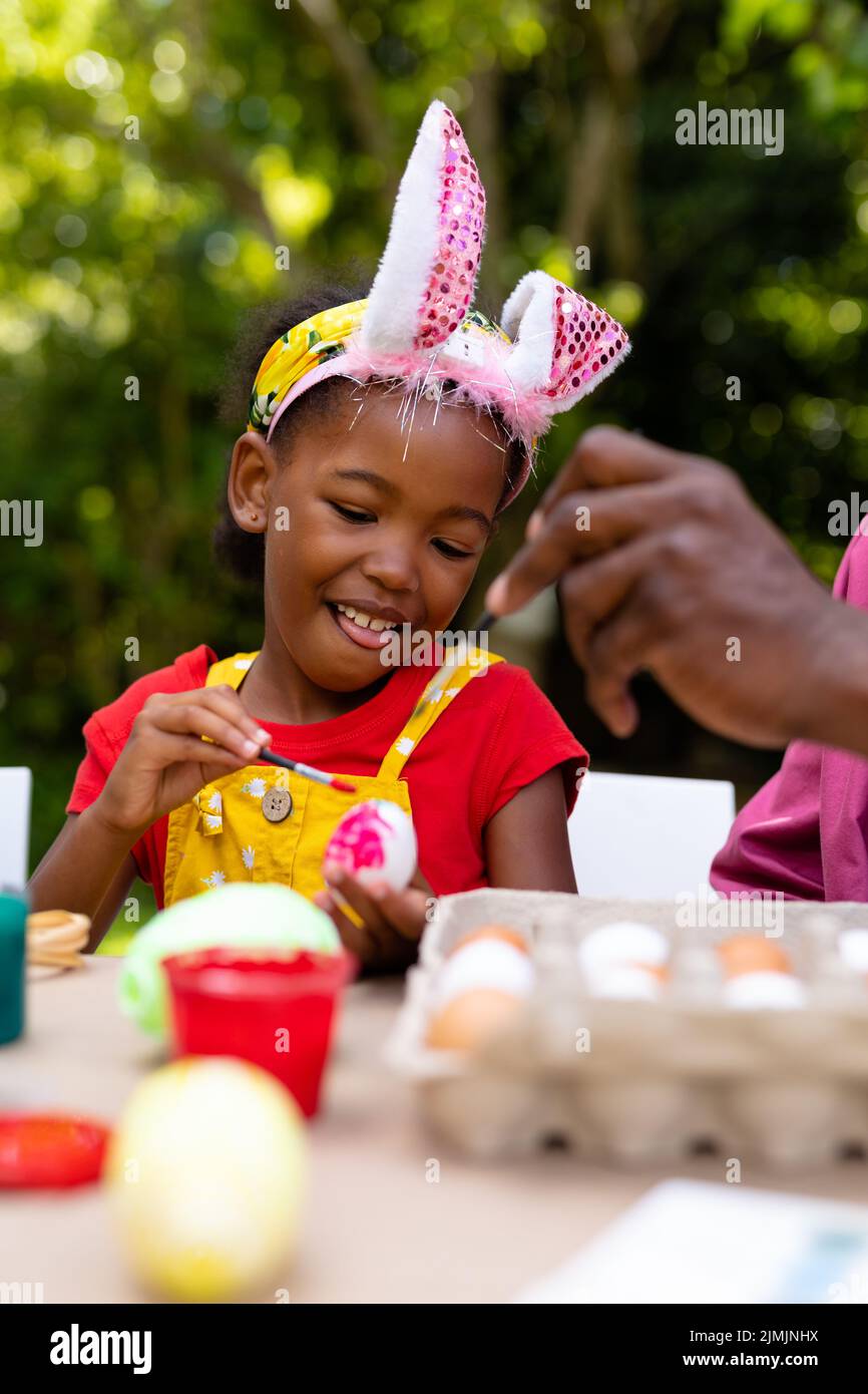 Smiling african american girl and grandfather in bunny ears painting egg in backyard on easter day Stock Photo