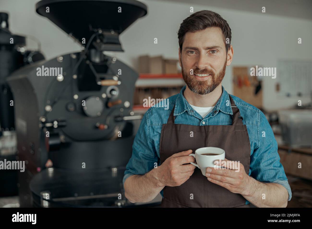 Smiling business owner with cup of fresh coffee on background of roasting machine Stock Photo