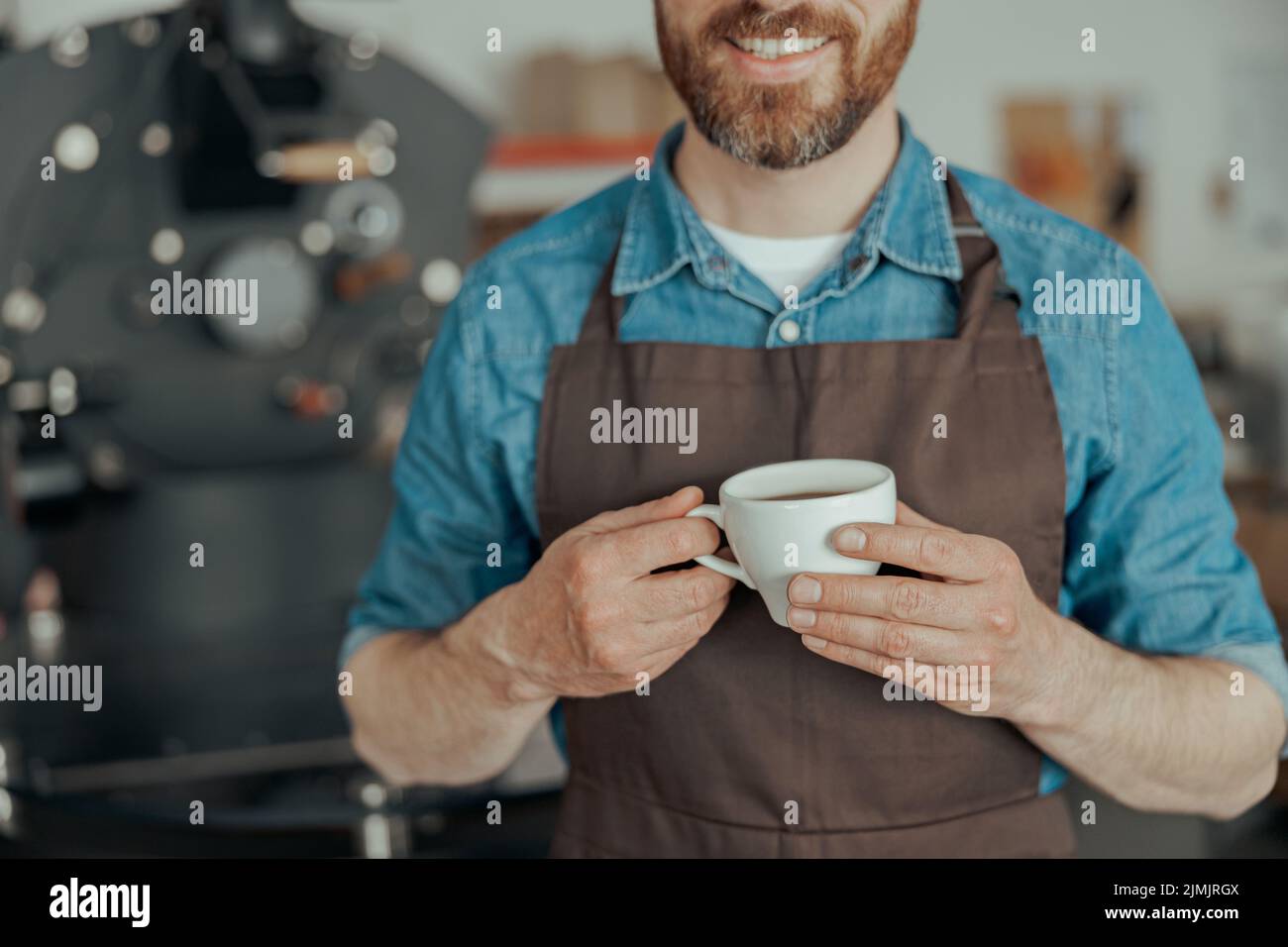 Close up of smiling business owner with cup of fresh coffee on background of roasting machine Stock Photo