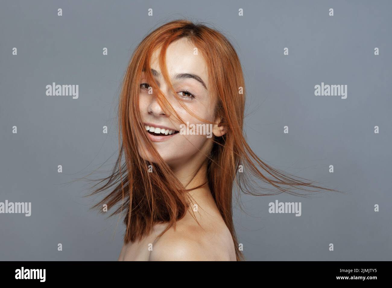 Beautiful dark burnt orange windy hair girl smiling. Studio portrait with happy face expression against gray background. Stock Photo
