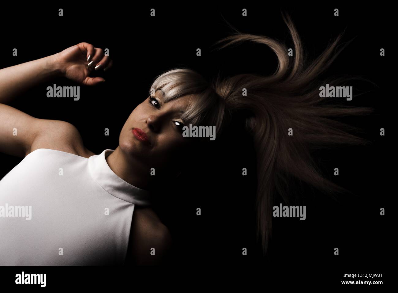 Beautiful girl with blond bangs fringe wearing white fashionable fancy dress lying against black background. Stock Photo