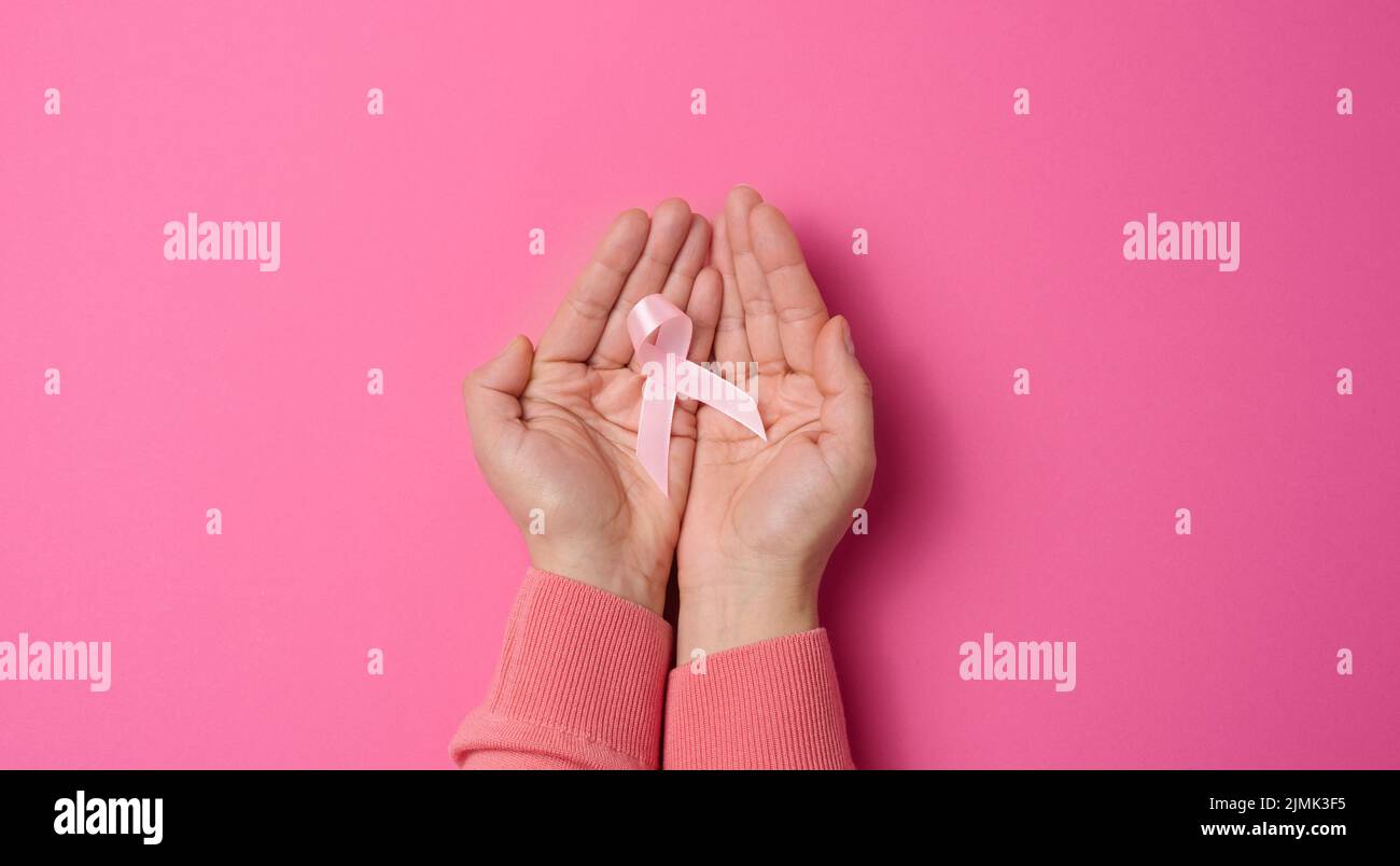 Two female hands hold a pink silk ribbon in the form of a loop on a pink background. Symbol of the fight against breast cancer Stock Photo