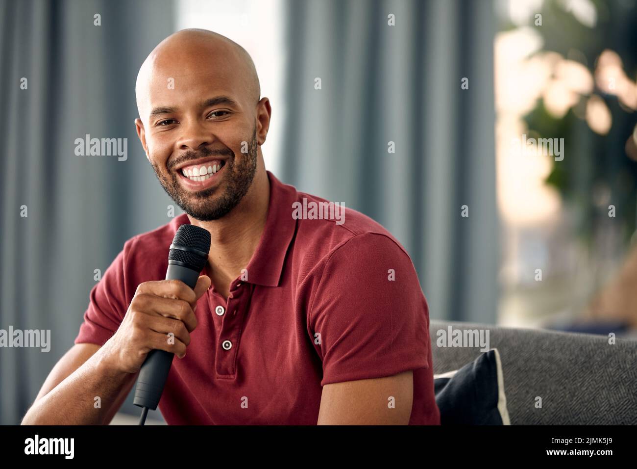Hes quite the talker. a young man sitting on a sofa while speaking over a microphone. Stock Photo