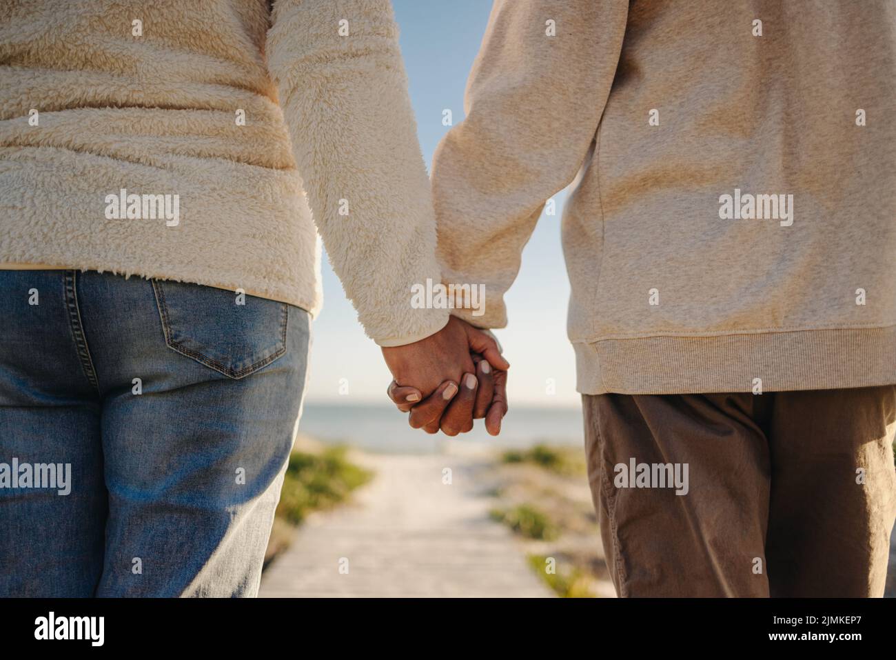 Romantic senior couple holding hands while walking down a boardwalk at the beach. Unrecognizable elderly couple taking a refreshing seaside holiday af Stock Photo
