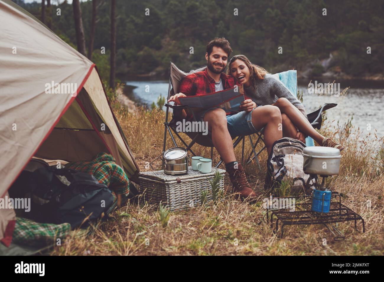 Loving young couple reading a travel map while sitting together outside their tent. Happy young couple camping next to the lake. Stock Photo