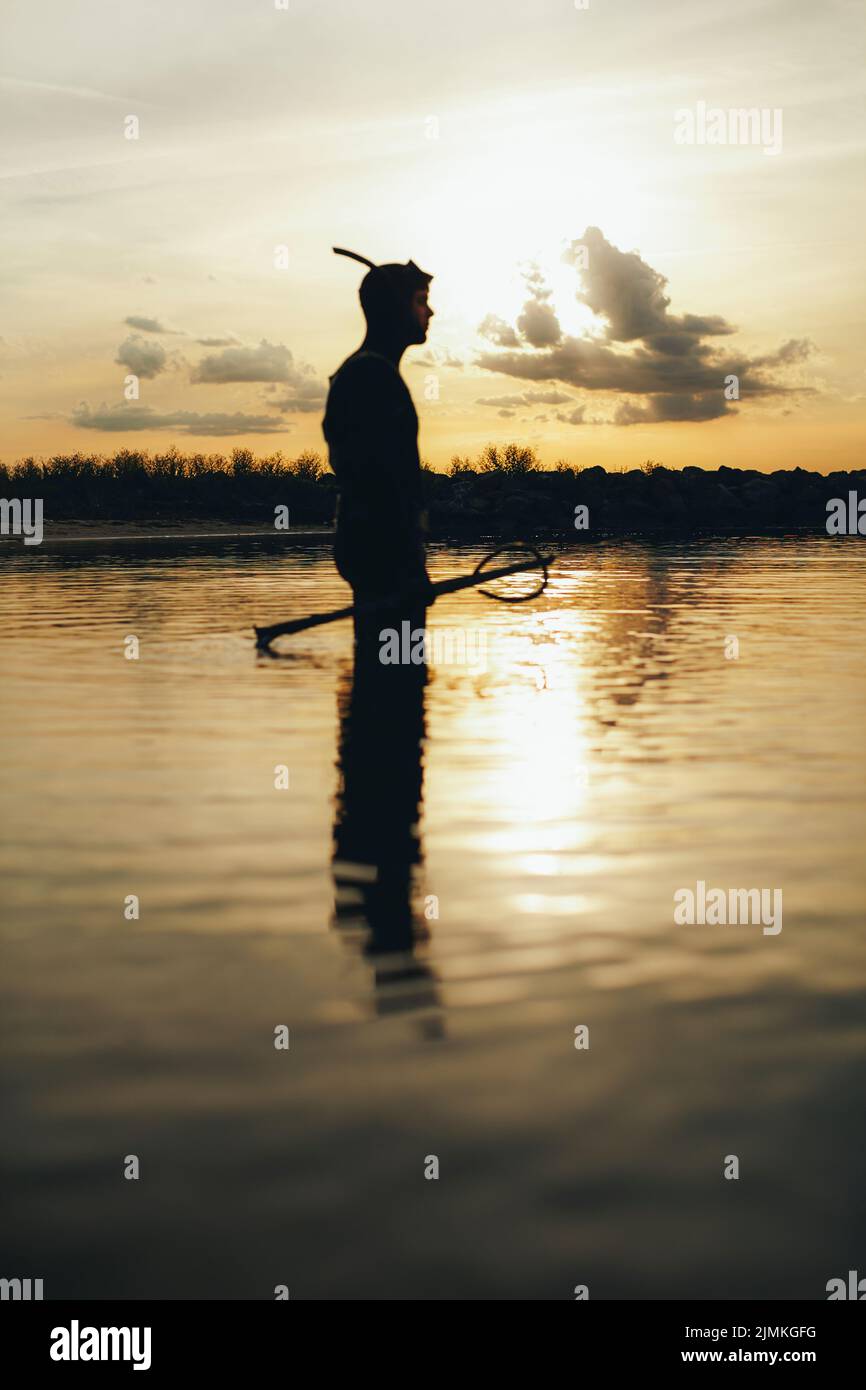 Side view of a male diver standing in sea water with a speargun. Adventurous young man going spearfishing at sunset. Stock Photo