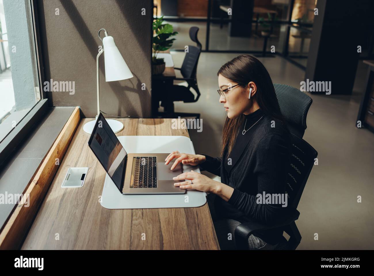 Focused young businesswoman using a laptop and wireless earphones while working alone. Young female entrepreneur working remotely in a modern co-worki Stock Photo
