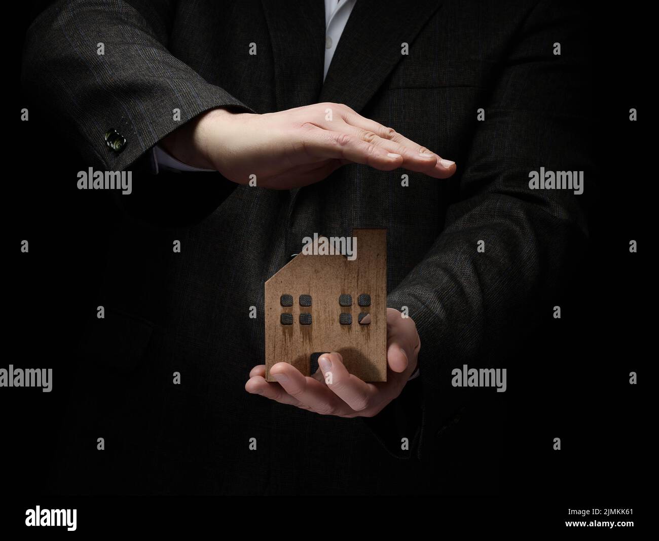 Businessman in a suit holds a wooden model of a house on a dark background. Real estate sale and purchase concept, rent. Insuran Stock Photo