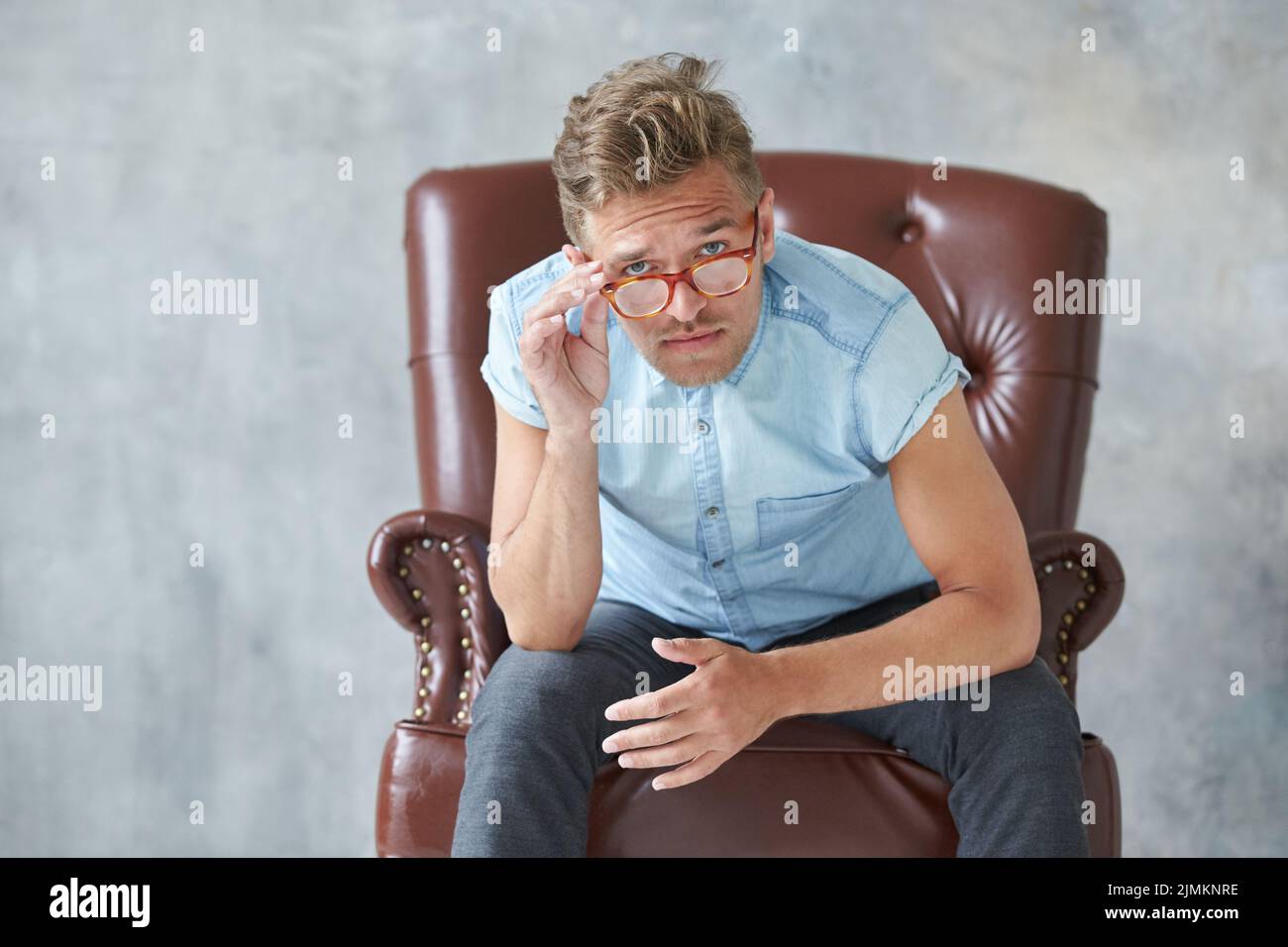 Portrait of a stylish intelligent man stares into the camera, small unshaven, charismatic, blue shirt, sitting on a brown leathe Stock Photo