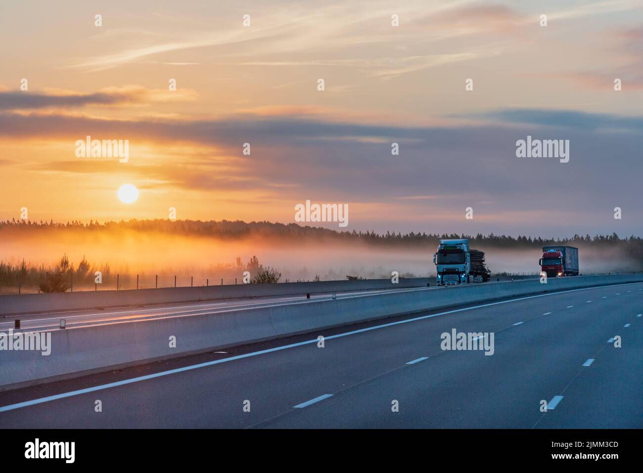 Landscape of a highway at dawn with the sun beginning to rise, two trucks circulating and the mist tucked between the trees. Stock Photo