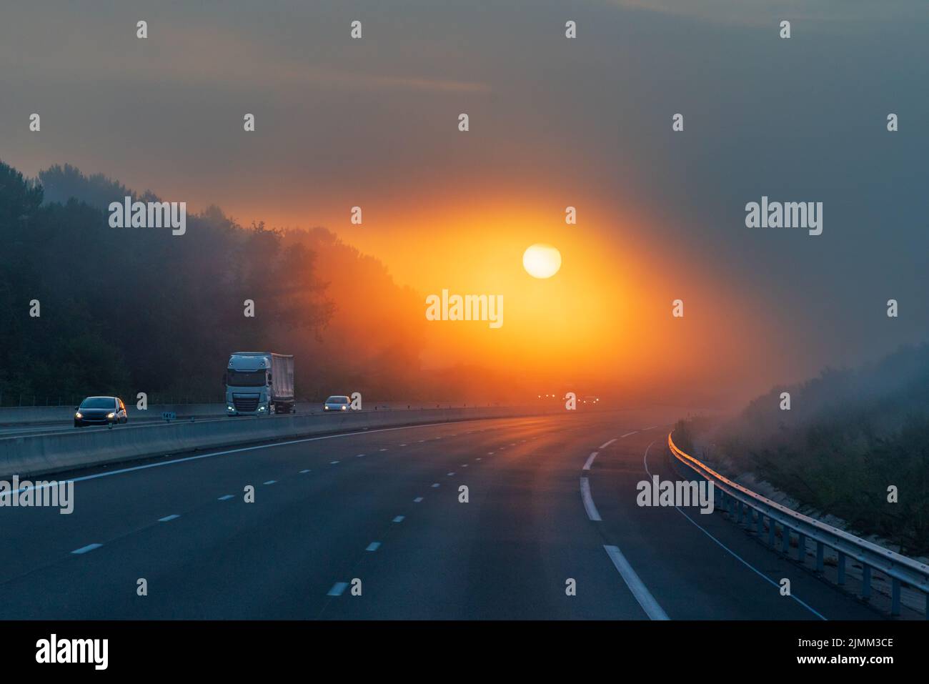 Landscape of a highway with a truck and several vehicles circulating at dawn, and the sun appearing through the clouds, allowing its circumference to Stock Photo
