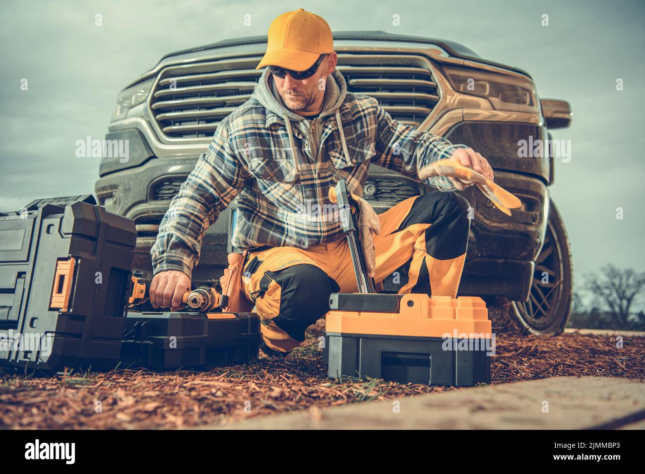 Caucasian Male Contractor Kneeling by Toolboxes Just Taken Out of His Pickup Truck Preparing the Necessary Tools for the Full Day of Work at the Build Stock Photo