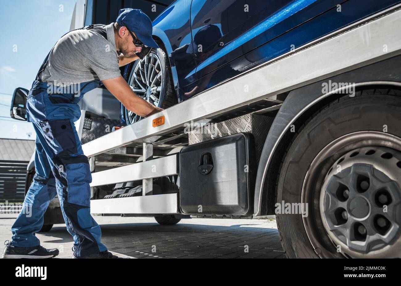Towing Worker Delivering Brand New Car from Dealer to Client. Caucasian Man Checking the Fastenings to Ensure Safe Transportation on His Tow Truck. Stock Photo