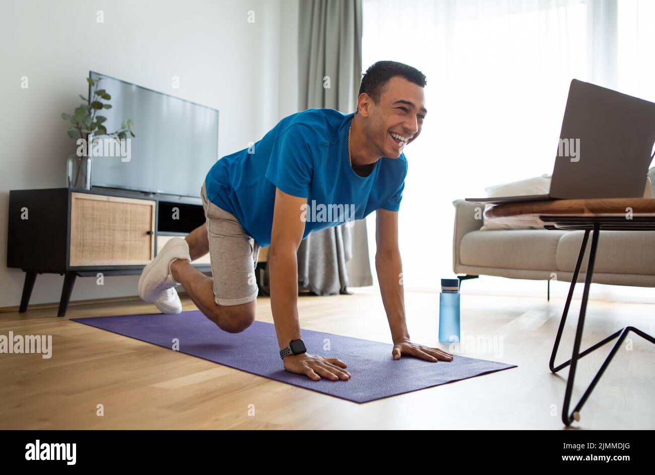 Active arab man in sportswear exercising at home, working on abs and arms strength, looking at laptop screen Stock Photo