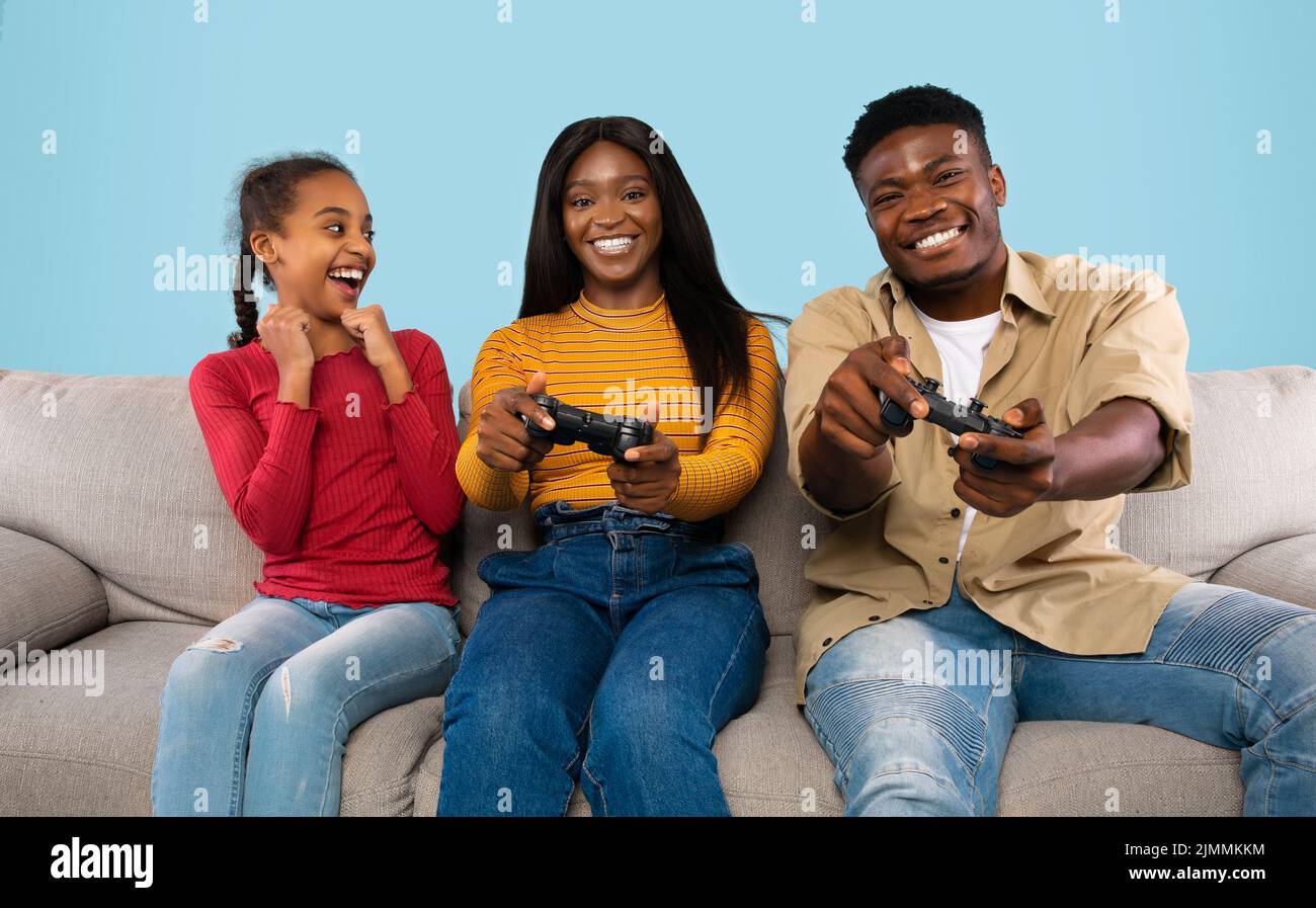 Family leisure. Joyful black girl cheering while parents playing videogames, sitting on sofa over blue background Stock Photo