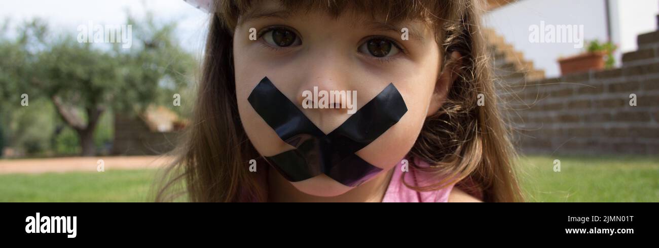 Image of an adorable little girl with a sad expression who has her mouth taped shut. Horizontal banner Stock Photo