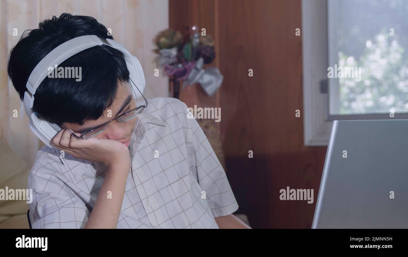 Bored sleepy student wearing headphone tired of learning doing homework sitting half a sleep at laptop screen in his room. Sad teenager boy studying p Stock Photo