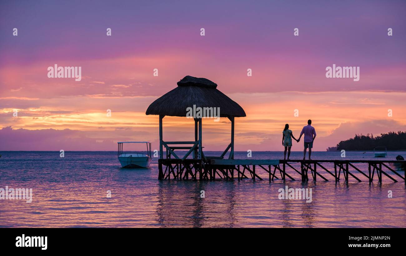 Couple watching sunset on a tropical beach with wooden pier in the ocean, men and woman sunset on the beach during luxury vacati Stock Photo