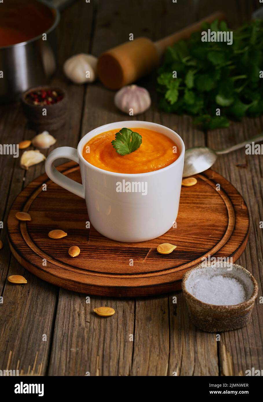 Pupmkin cream soup in cup on brown wooden table, vertical, side view. Dietary vegetarian puree on cutting board with parsley, ga Stock Photo