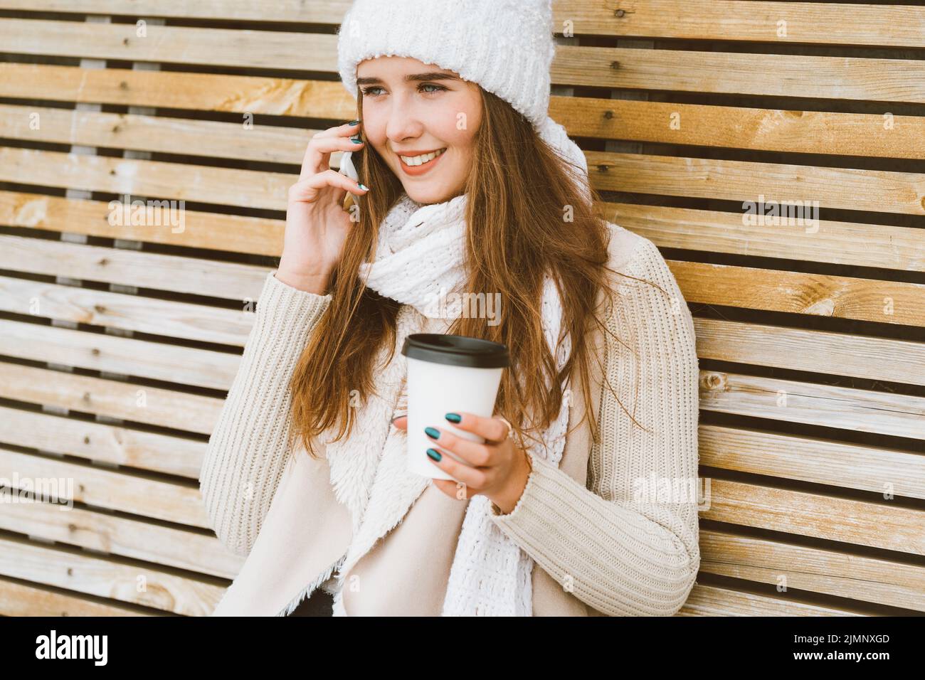 Beautiful young girl drinking coffee, tea from plastic mug in autumn, winter and talking on mobile phone. Woman with long hair s Stock Photo