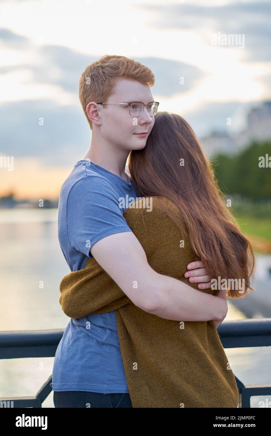 Girl with long thick dark hear embracing redhead boy in the blue t-shirt on bridge, teen love at sunset Stock Photo