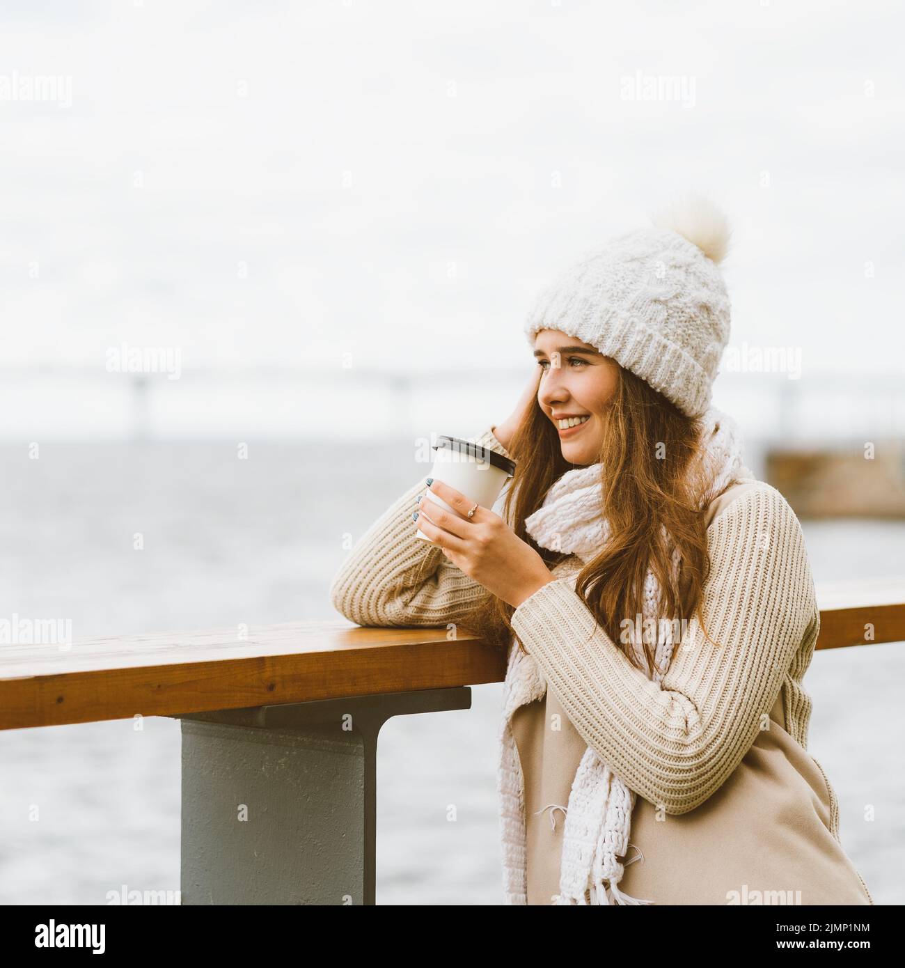 Beautiful young girl drinking coffee, tea from plastic mug in autumn, winter. A woman with long hair stands on waterfront on Bal Stock Photo