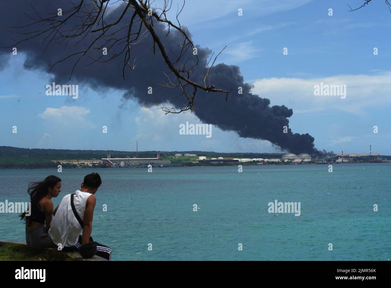 A couple sits near the sea as smoke rises over fuel storage tanks that