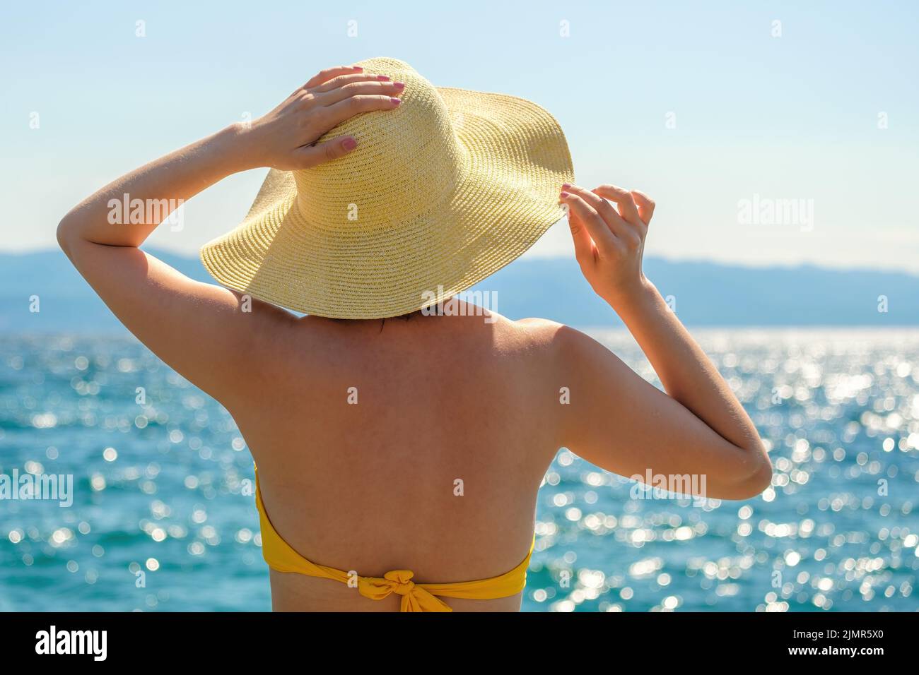 Elegant lady in yellow bikini admires sparkling sea at resort at sunlight. Young woman holds straw hat with hands on windy summer day close back view Stock Photo
