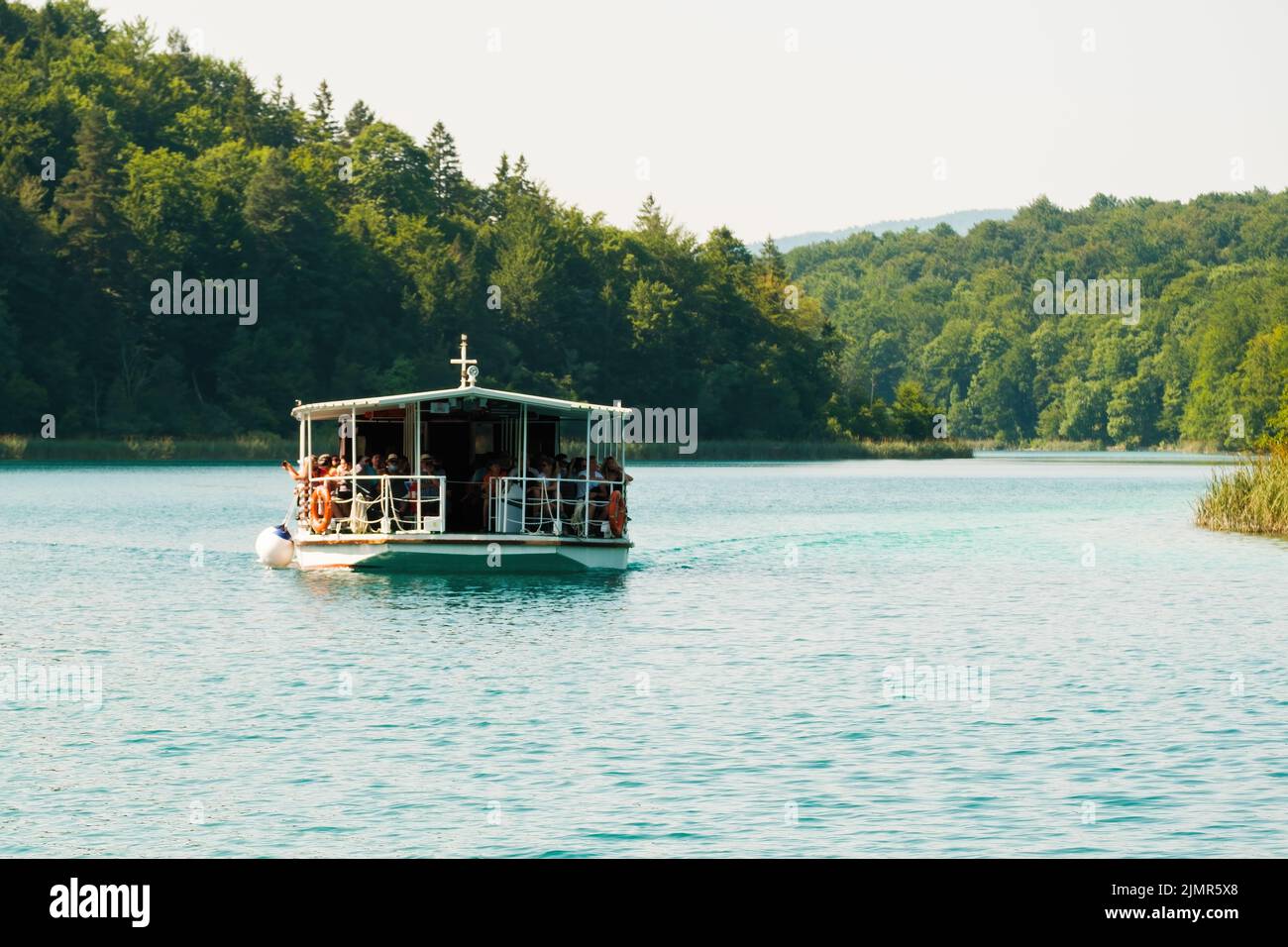 Boat with group of tourists sails on deep turquoise lake surrounded by green forestry hills. Ship excursion on blue lagoon in natural park reserve on Plitvice lakes Stock Photo