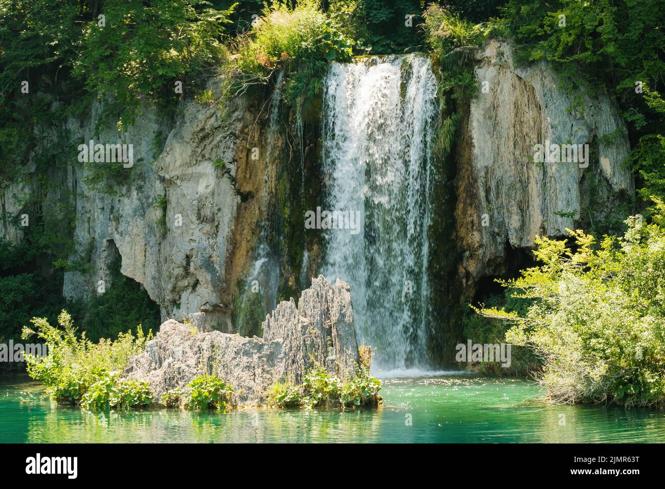 Waterfall flows down from rocky cliff into clear turquoise water at sunlight. Breathtaking view of aquamarine cascade in national park on Plitvice lakes Stock Photo