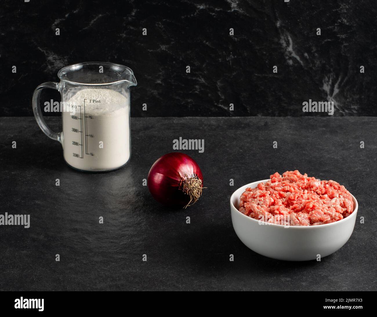 Plate with fresh ground beef, red onion, measuring cup with flour on a grey black background, space for text. Stock Photo