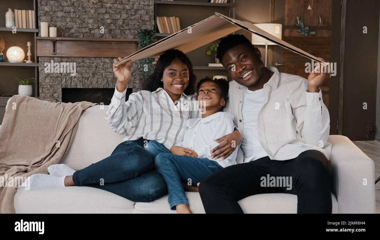 African couple sitting on couch under cardboard roof smiling looking at camera active happy daughter girl running sits on sofa. Relocation day to new Stock Photo