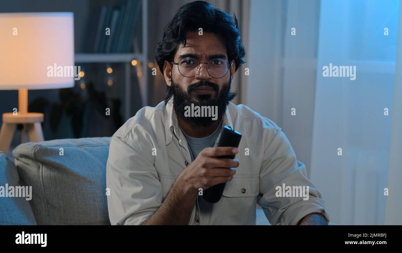 Evening entertainment Arabian Indian man bearded male in glasses in darkness living at home guy sits sofa switch change channel choosing program movie Stock Photo