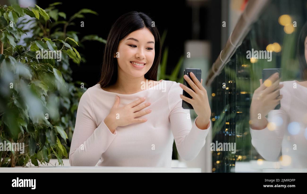 Asian korean friendle girl japanese lady chinese female smiling waving hello to phone webcam woman with mobile gadget talking make video call in cafe Stock Photo