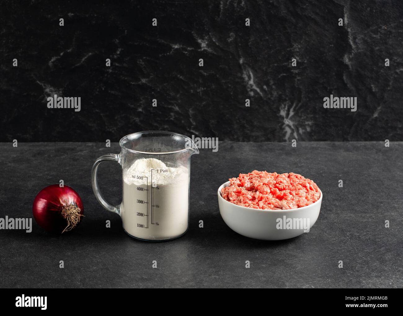 Plate with fresh ground beef, red onion, measuring cup with flour on a grey black background, space for text. Stock Photo