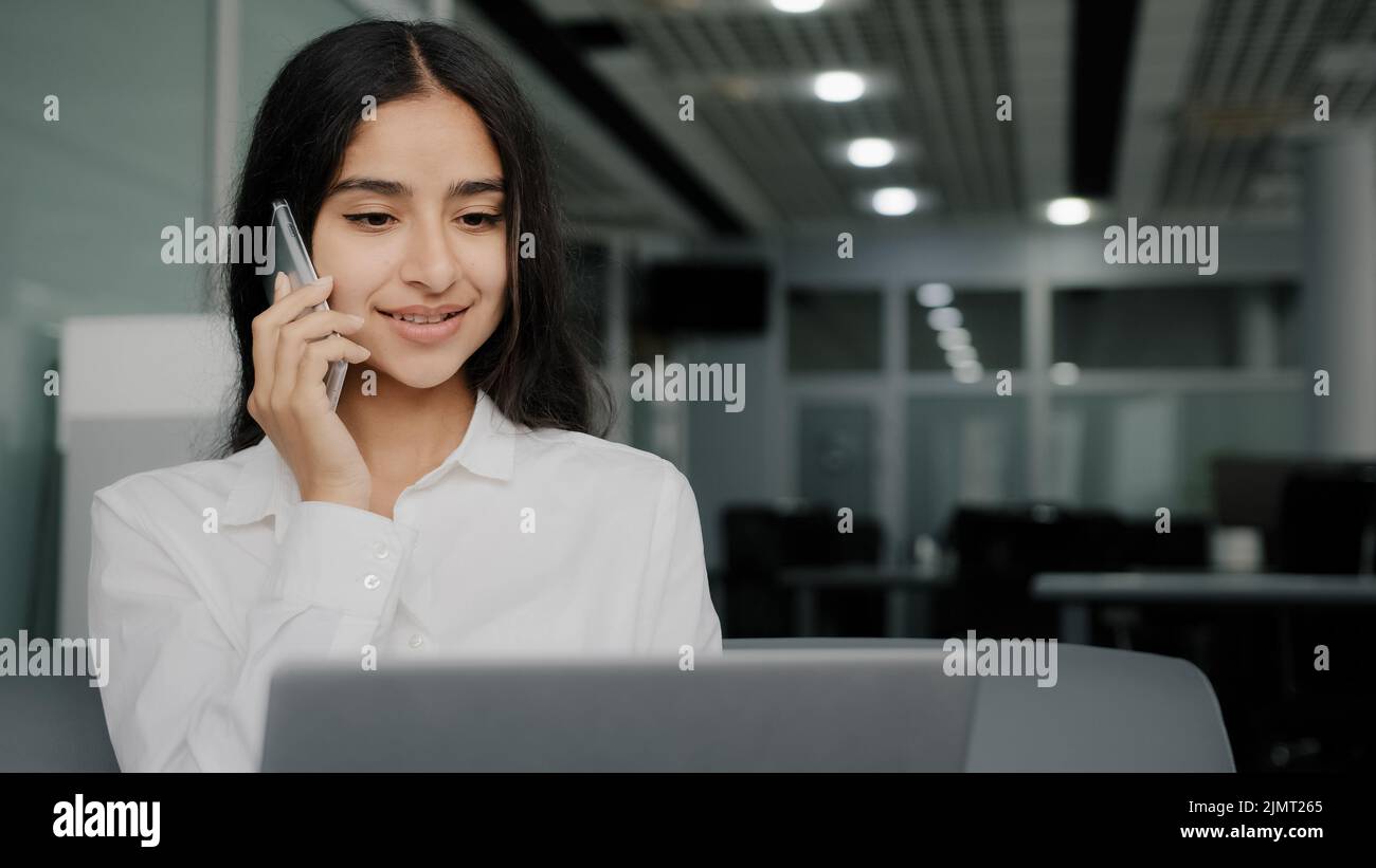 Busy female worker woman talk phone answer call arabic businesswoman do multitasking work on laptop chatting provide mobile support assistance for Stock Photo