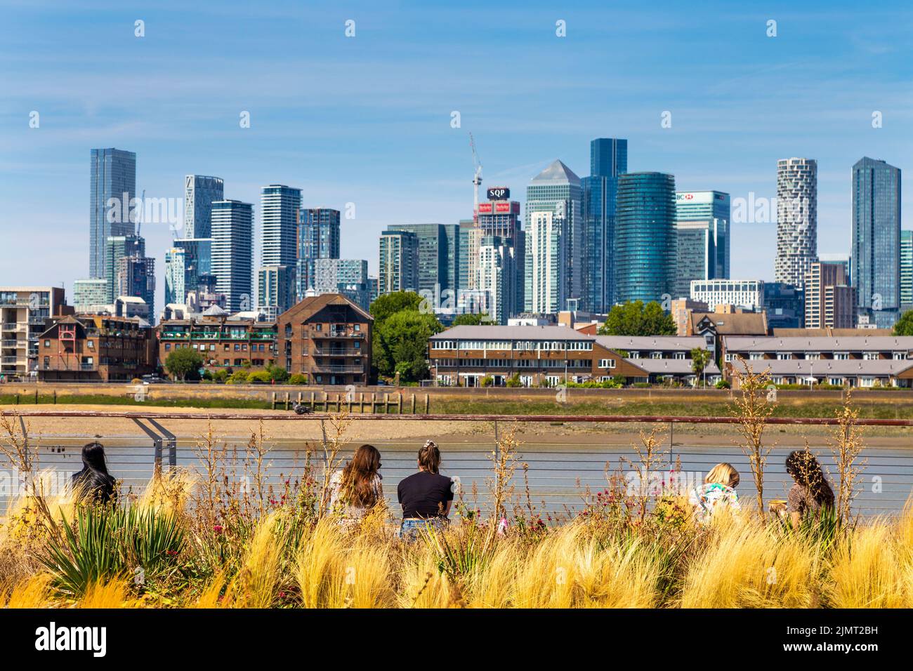 People sitting along Greenwich riverside overlooking Canary Wharf skyscrapers, London, UK Stock Photo