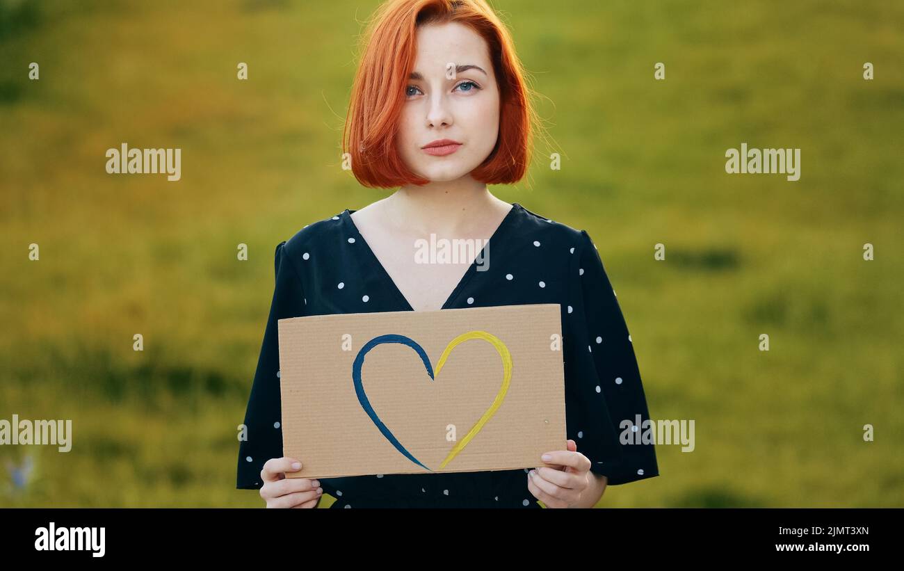 Unhappy sad redhead woman hold artwork colorful cardboard drawing blue yellow love shape heart romantic symbol demonstrate tenderness feelings Stock Photo