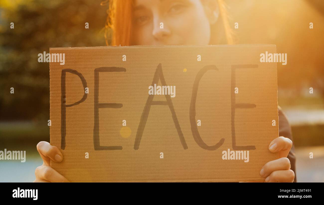 Close up female hands hold cardboard with written word peace symbolic banner attractive young woman pretty redhead girl stand in park demonstrate Stock Photo