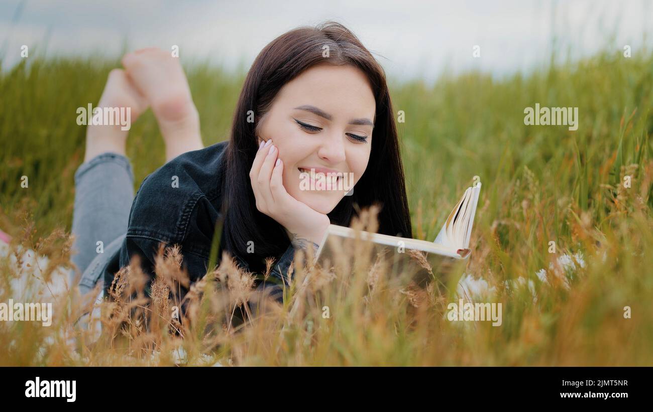 Young serene caucasian brunet woman girl lying on grass outdoors attractive smiling teenage student reads book enjoy romantic literature history relax Stock Photo