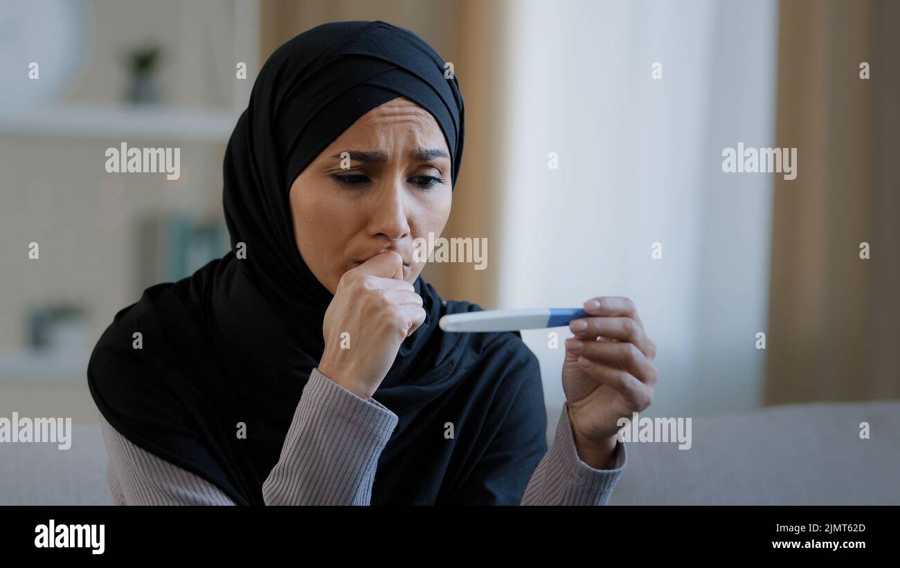 Shocked serious islamic young arabian woman muslim girl wear hijab female adult pregnant sit at home feeling fear look at positive pregnancy test Stock Photo