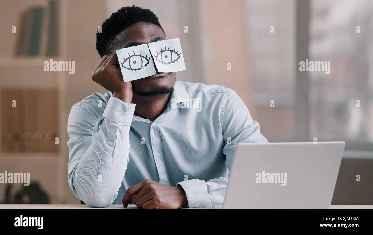 Sleepy funny african american office manager business man sitting at home workplace with sticky notes on eyes comic young tired worker has sleeping pr Stock Photo