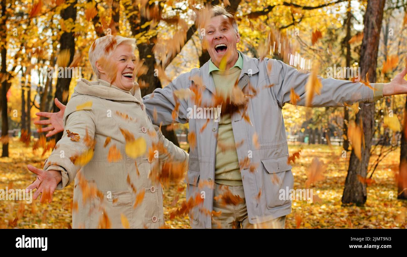 Happy married elderly caucasian couple partners in civil park mature woman senior man scatter leafage outdoors throw up autumn fallen leaves lovely pe Stock Photo