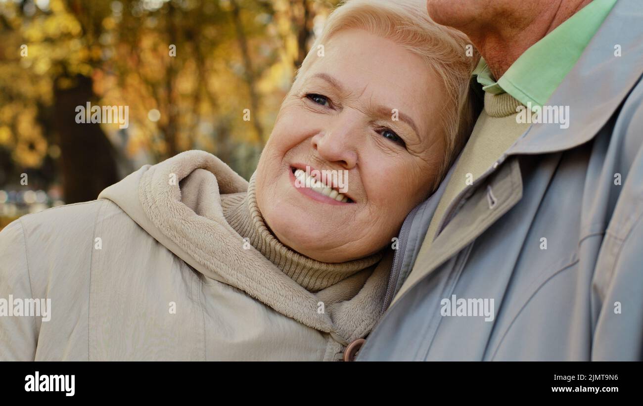 Happy middle aged pretty caucasian elderly woman grandma smiling stand relax in autumn park spend time outdoors lean on shoulder mature partner man Stock Photo