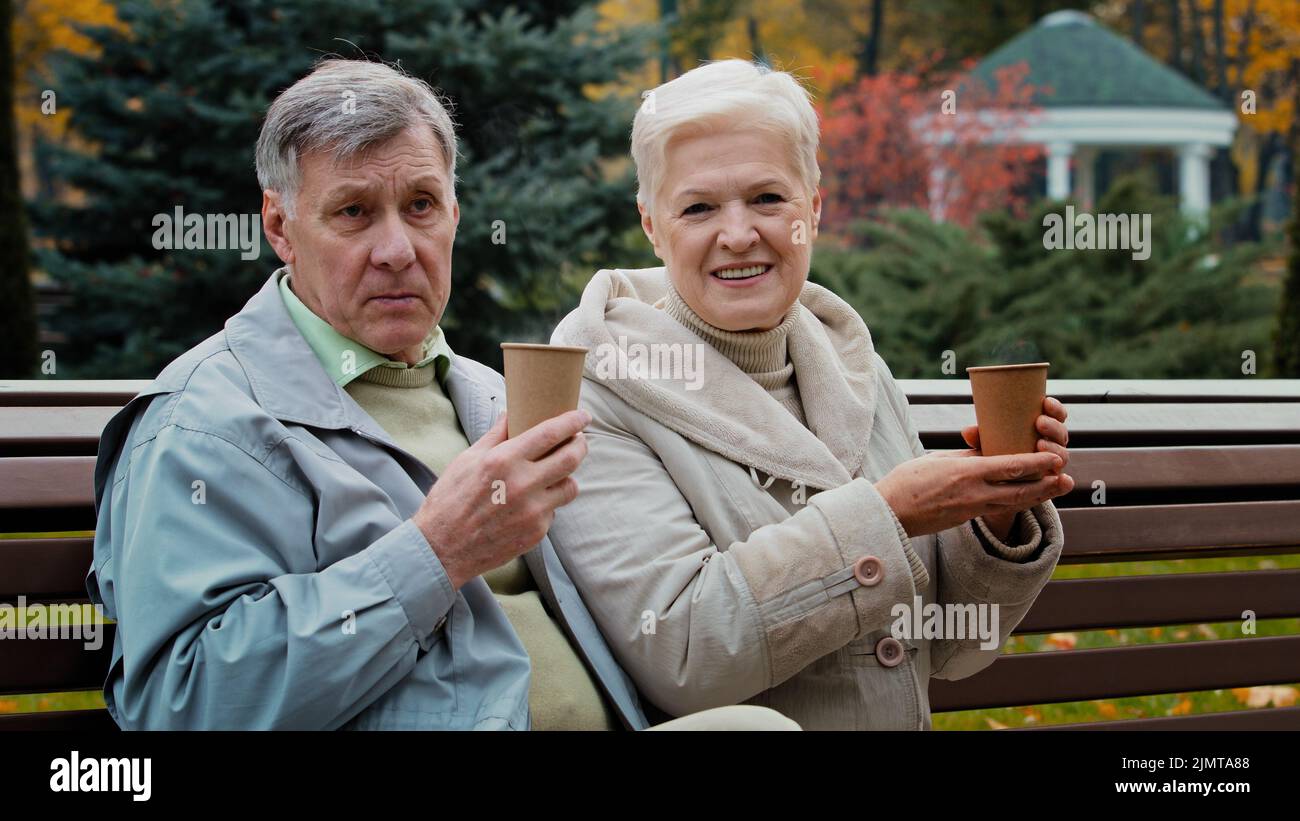 Carefree elderly grandparents drinking hot coffee tea outdoors senior aged couple man woman pensioners grandparents family sit on bench enjoy Stock Photo
