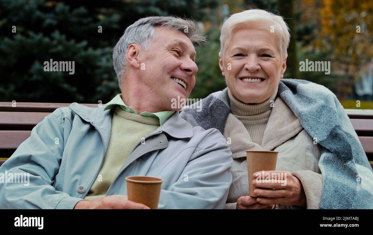 Happy senior aged couple talking sit in autumn park rest together carefree elderly grandparents drinking coffee tea outdoors enjoying pleasant convers Stock Photo