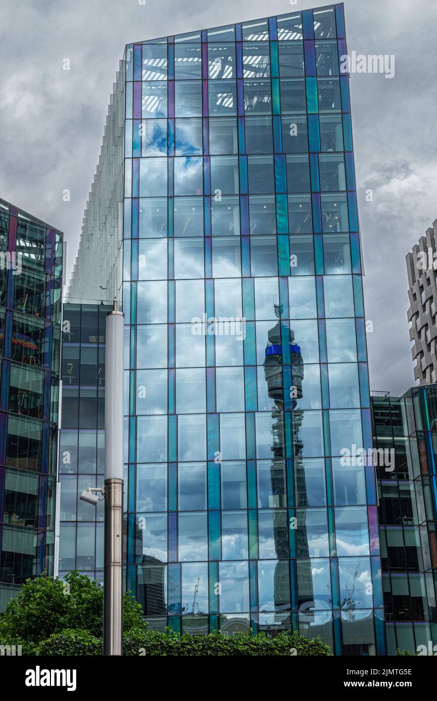 London, Great Britain - July 3, 2022: Tall slender BT tower reflected in blueish glass facade of Regent's Place office building. Green foliage at bott Stock Photo